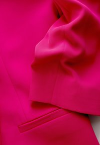 Fuchsiová látka s textúrovaným, rebrovaným povrchom; obsahuje hladké švy a malý detail vrecka, ktorý zdôrazňuje jemnosť a plynulosť.