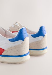 Fehér tornacipők piros és kék díszítésekkel, sima bőr és velúr anyagból, ívelt kialakításúak, texturált sarokkal és gumitalppal.