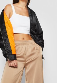 Veste bomber en satin noir avec un détail de manche orange, associée à un top court blanc et un pantalon en satin beige avec taille élastique.