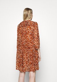 Femme aux longs cheveux blonds portant une robe ample, mi-longue, orange et marron à motifs, vue de dos sur un fond blanc uni.