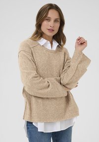 Beiger Wollmischpullover mit lockerer Passform, weiten Ärmeln und rundem Ausschnitt, über einem weißen kragenlosen Hemd getragen.