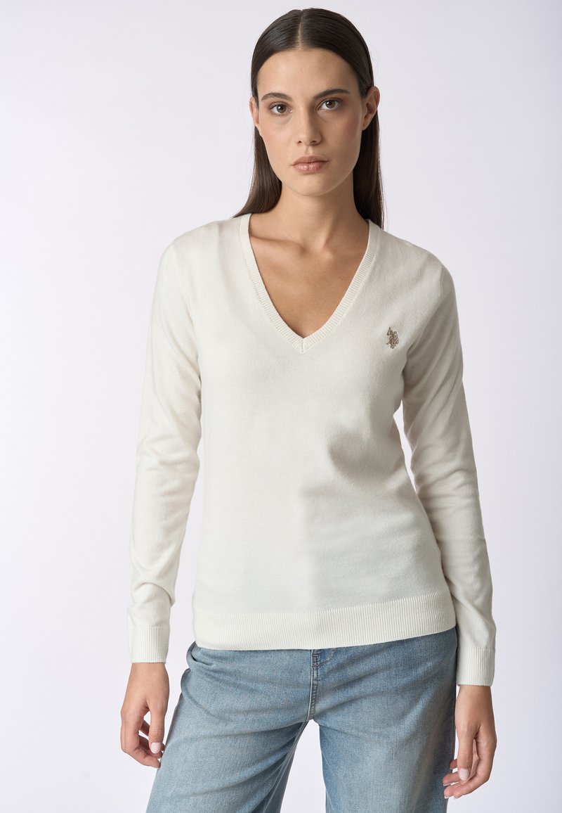 Maglione crema a V in tessuto morbido e leggero. Presenta maniche lunghe e polsini a costine, con un piccolo logo ricamato sul petto.