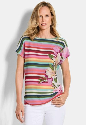 Frau trägt ein mehrfarbiges, horizontal gestreiftes Hemd mit großem pinkfarbenem Blumenmuster auf einer Seite, kombiniert mit weißen Hosen.