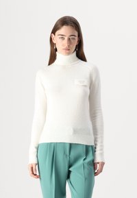 Maglione bianco a collo alto con una finitura morbida e strutturata, caratterizzato da una piccola patch con logo. Abbinato a pantaloni teal, con un design aderente.