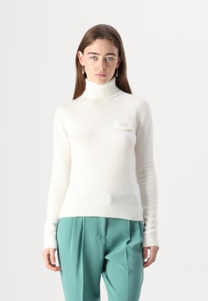 Maglione bianco a collo alto con una finitura morbida e strutturata, caratterizzato da una piccola patch con logo. Abbinato a pantaloni teal, con un design aderente.