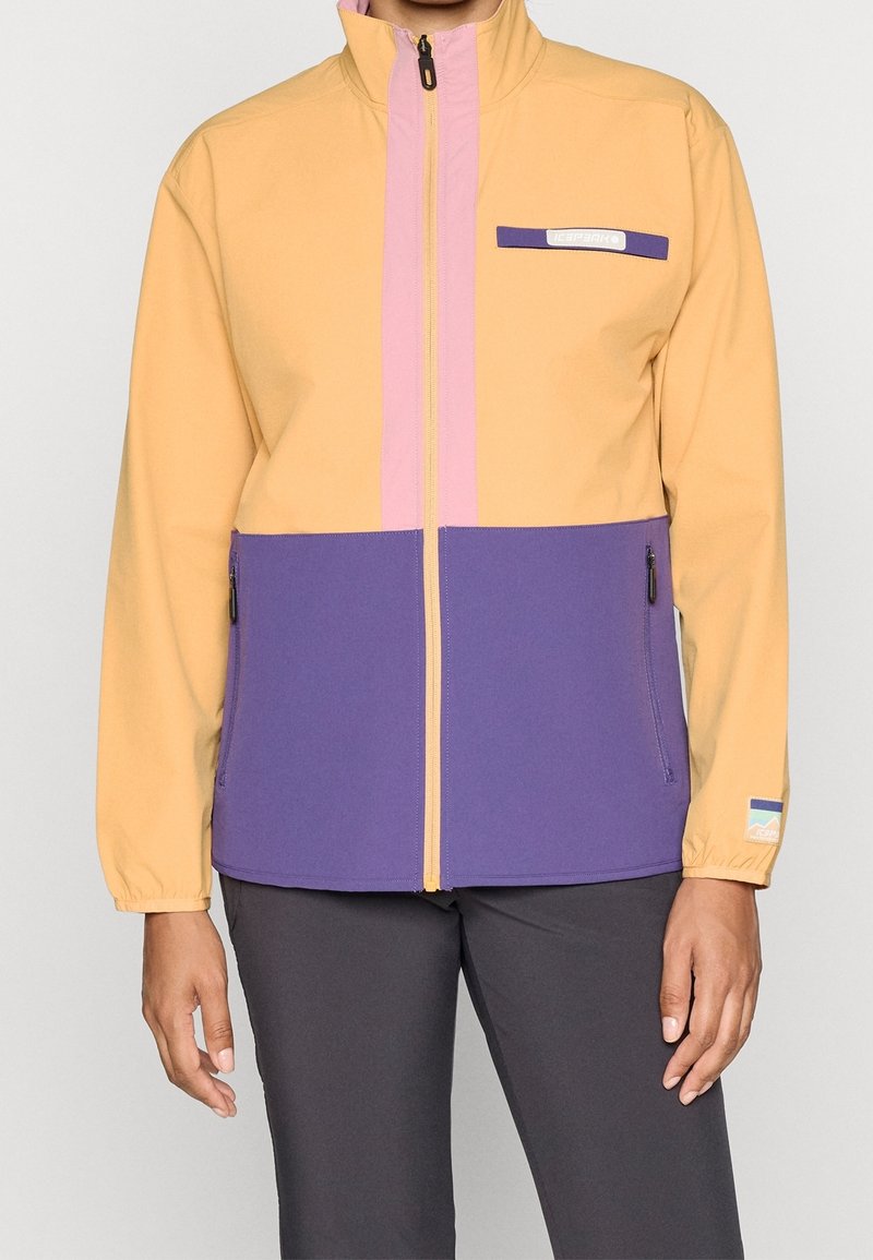 Personne portant une veste zippée avec le haut jaune, le bas violet et une bande rose au centre, associée à un pantalon foncé.