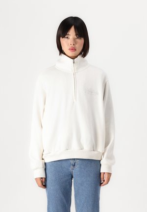 Sweatshirt blanc zippé avec un col haut, présentant une finition texturée et un logo brodé. Associé à un jean en denim bleu clair.