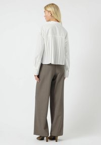 Camicia bianca con schiena plissettata, abbinata a pantaloni larghi grigio scuro e tacchi marroni. La camicia presenta un colletto classico e polsini con bottoni.