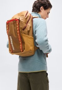 Άτομο που φοράει ανοιχτό μπλε φλις μπουφάν και καφέ παντελόνι, κουβαλώντας μεγάλο σακίδιο πλάτης της Patagonia σε χρώμα μουσταρδί και καφέ με κόκκινες λεπτομέρειες.