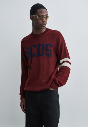 Maglione a maglia marrone con lettering navy "GCDS", con accenti bianchi a righe sulle maniche. Indossato con pantaloni neri e occhiali moderni.