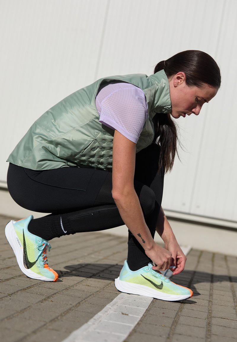 Mujer vestida con ropa deportiva atándose los zapatos de correr en neón azul y naranja en un camino pavimentado frente a un fondo de pared blanca.