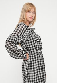 Robe à carreaux noir et blanc en tissu léger, avec de longues manches volantes, une taille cintrée et un détail à nouer à la taille.
