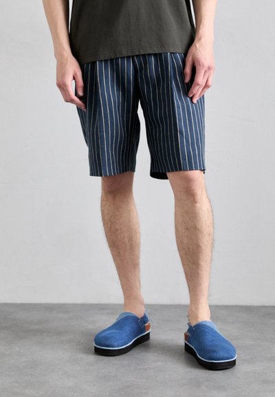 Blaue und weiße gestreifte Shorts aus leichtem Material, kombiniert mit blauen Clogs mit Holzabsatz und einem Riemen hinten.
