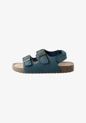 Sandal din suede teal cu două curele ajustabile și catarame din alamă, având o branț texturat bej și o talpă din cauciuc maro.