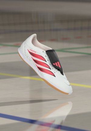 Chaussure de football indoor blanche avec des bandes rouges, une semelle en gomme et une protection noire sur les lacets, flottant au-dessus d’un sol de gym avec des marquages colorés du terrain.