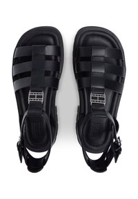 Μαύρα λαστιχένια σανδάλια με σχέδιο λουριών, ρυθμιζόμενο λουρί στον αστράγαλο και μεταλλικά στοιχεία με το λογότυπο μπροστά. Ομαλή υφή με επίπεδες σόλες.