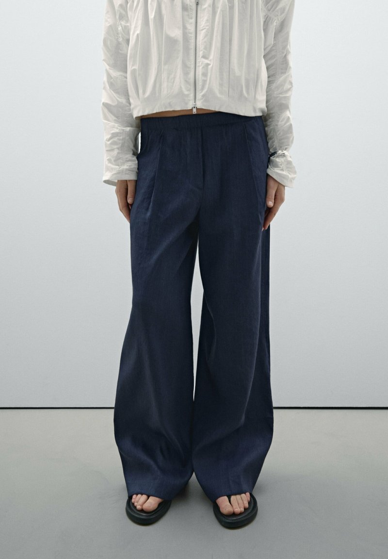 Veste blanche courte zippée à l'avant, pantalon large bleu marine et sandales noires à bout ouvert sur une personne debout devant un fond uni.