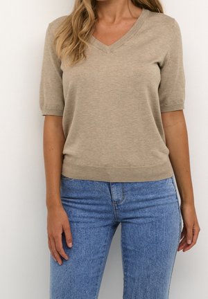 Femme portant un pull beige à col en V et manches courtes, ainsi qu'un jean bleu, debout devant un mur blanc uni.