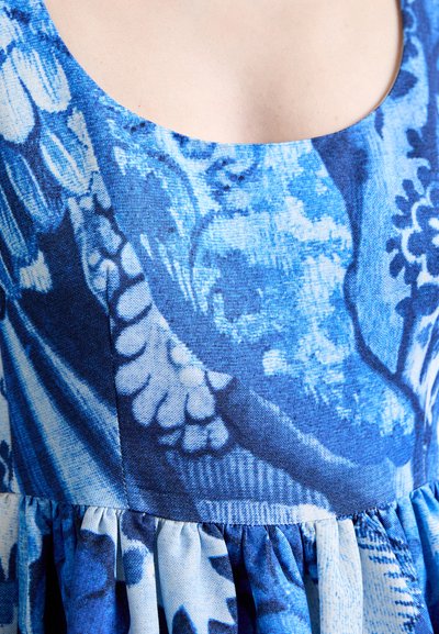 Blue patterned fabric with floral and abstract motifs. Features a scoop neckline and a gathered waist design. Textured, lightweight material.