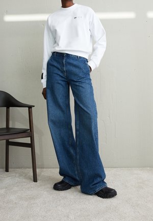 Osoba v bílém dlouhém tričku, širokých modrých džínách a černých robustních botách stojí vedle černé dřevěné židle na světlém koberci.