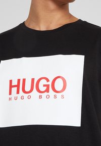 Sort bomulds T-shirt med et stort hvidt rektangulært grafik med rød tekst "HUGO" og "HUGO BOSS" stiliseret nedenunder. Klassisk crew neck design.