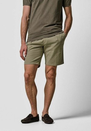 Olivegrüne Herrenshorts mit Seitentaschen, mit tailliertem Schnitt und glattem Baumwollstoff. Kombiniert mit einem passenden braunen T-Shirt und dunklen Loafers.