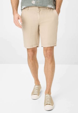 Beige Leinen-Shorts mit gerader Passform, ausgestattet mit Seitentaschen. Kombiniert mit hellen Schnürsneakern und einem gemusterten Hemd.