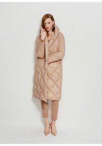 Kobieta ubrana w długi beżowy pikowany płaszcz z wysokim kołnierzem, zestawiony z podwiniętymi wąskimi spodniami i czółenkami z ostrym noskiem, stojąca przy białej ścianie.