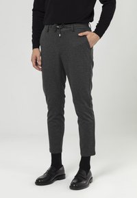 Graue, schmal zulaufende Hose mit einer Kordelzugtaille. Figurbetontes Design, glatte Textur, Seiten Taschen, kombiniert mit schwarzen Slippern und Socken.