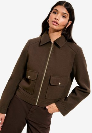 Mujer que lleva una chaqueta con cremallera de color marrón oscuro, con dos bolsillos con solapa en el frente y pantalones marrones, de pie contra un fondo blanco.