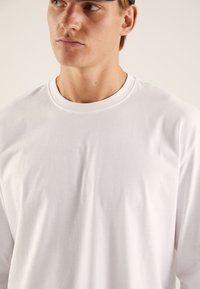 Langärmliges weißes T-Shirt aus weicher Baumwolle, mit rundem Halsausschnitt und glatter Textur, kaum knitternd. Einfaches Design, ohne Muster.