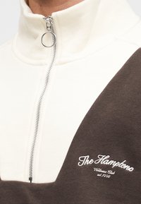 Φούτερ με φερμουάρ στο μισό μήκος, με άσπρο πάνω μέρος και καφέ κάτω μέρος, με κεντημένο κείμενο και μεταλλικό φερμουάρ με κρίκο στο τράβηγμα.