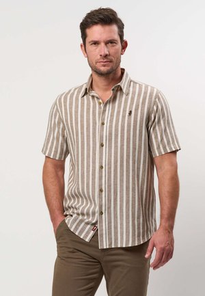 Mies, jolla on lyhythihainen ruskea- ja valkoraidallinen nappipaita ja khakihousut, seisoo yhdellä kädellä taskussa.