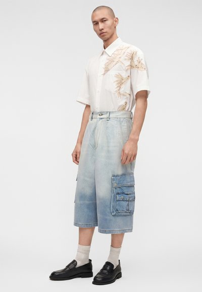 Άνδρας που φοράει λευκό πουκάμισο με κοντά μανίκια και μπεζ σχέδια φύλλων, ανοιχτό μπλε μακρύ τζιν cargo σορτς, μπεζ κάλτσες και μαύρα loafers.