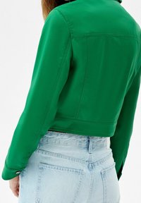 Giacca verde corta con una texture liscia e lucida. Presenta un colletto classico e polsini con bottone, abbinata a jeans di denim azzurro chiaro.