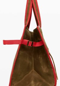 Canvas tote bag in brown with red accents. Features a collapsible design and adjustable strap with buckle. Textured surface, open top.