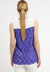 Top senza maniche con una parte superiore bianca e una parte inferiore in pizzo blu con un motivo floreale. Tessuto liscio e design aderente.