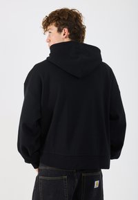 Sudadera negra con capucha grande, hombros caídos y dobladillo acanalado. La tela parece suave con una textura lisa.