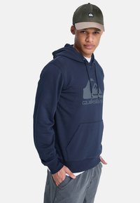 Sudadera con capucha de color azul marino con un bolsillo delantero. Presenta un logo gris, tejido suave y puños acanalados. Lleva puesta con pantalones grises y una gorra.