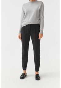 Maglione grigio chiaro a maniche lunghe con scollo a girocollo, abbinato a pantaloni neri su misura con gamba affusolata e dettagli a piega. Mocassini neri.