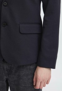 Blazer bleu marine à finition texturée, doté de deux boutons, d'une poche poitrine unique et de manches élégantes à coupe droite. Porté avec un pantalon gris foncé.