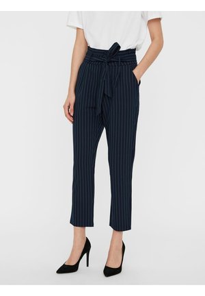 Ołówkowe granatowe spodnie w drobną prążki z wysokim stanem i związanym paskiem, połączone z białą bluzką i czarnymi butami na wysokim obcasie, noszone przez stojącą osobę.