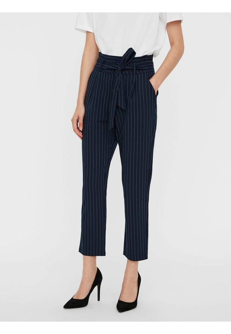 Pantalon à fines rayures bleu marine taille haute avec ceinture nouée, associé à un haut blanc et des chaussures à talons noirs, porté par une personne debout.
