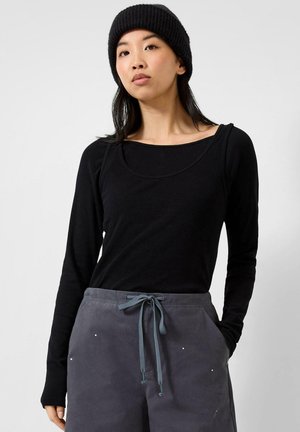 Junge Frau trägt ein schwarzes Langarmshirt, eine schwarze gestrickte Mütze und graue Hose mit Kordelzug, posiert mit den Händen in den Taschen vor einem einfarbigen Hintergrund.