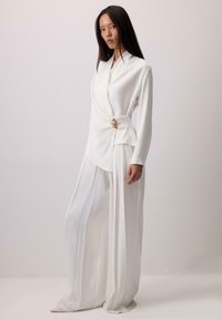 Bela majica z dolgimi rokavi z ovojnim dizajnom in zlato poudarkom, kombinirana z belimi širokimi hlačami z gubicami, ki ustvarjajo strukturni silhuet.