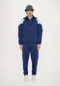 Marineblå atletisk jakke med lyseblå accenter, lynlås foran og elastiske manchetter, parret med matchende marineblå joggingbukser og hvide sneakers.