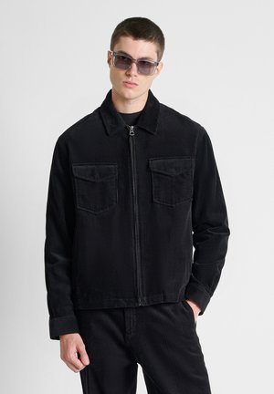 Jonge man draagt een zwart ribfluwelen jack en broek met een zonnebril tegen een effen witte achtergrond.