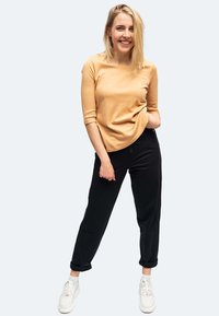 Junge Frau lächelt, steht mit einer Hand in der Tasche, trägt ein beiges Oberteil, schwarze Hose, weiße Socken und weiße Sneaker.