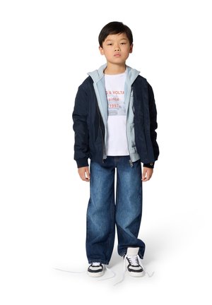 Veste aviateur marine avec un sweat à capuche bleu clair en dessous, t-shirt blanc à motif, jean bleu ample et baskets noir et blanc avec des lacets défaits.