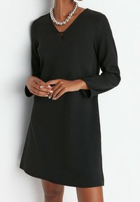 Kvinna iklädd en svart långärmad klänning med V-ringning och ett stort silverpärlhalsband, stående framför en vit bakgrund.